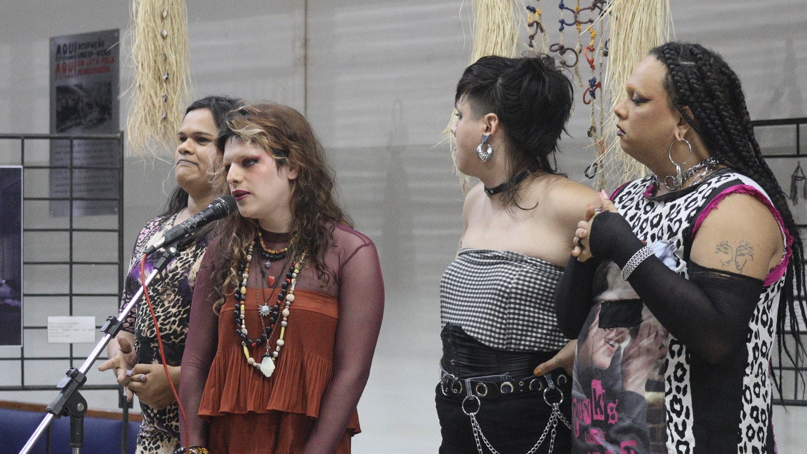 Fotografia. Quatro pessoas estão próximas a um microfone em pedestal. A pessoa à esquerda é negra de pele clara, tem cabelos lisos, pretos e longos, sobrancelhas espessas e lábios médios, usa uma blusa e calça com estampa de onça. A segunda é branca, tem cabelos ondulados castanhos com mechas loiras, usa uma blusa de tule manga longa vinho sobre outra terracota com detalhes de babados.  Fala ao microfone. Atrás, há uma pessoa branca de cabelos preto, curto e liso, raspado nas laterais, com estilo moicano. Usa uma blusa sem alças xadrez em preto e branco e calça preta, está com um cinto largo preto com argolas metálicas e correntes penduradas. Ao lado direito, uma pessoa negra de pele clara com tranças longas e grossas, usa uma blusa com tons de branco, preto e rosa. Está com uma luva longa preta.  Ao fundo, presas ao teto duas instalações com palha e búzios. 
