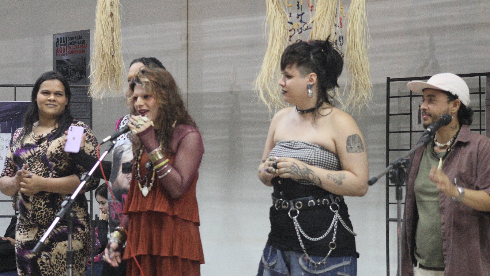  Fotografia. Cinco - pessoas próximas aos microfones. A da esquerda é alta, negra de pele clara, tem cabelos lisos, pretos e longos, sobrancelhas espessas e lábios médios. Está de blusa e calça com estampa de onça. Ao lado, uma pessoa com perfil de uma pessoa alta com tranças. A terceira pessoa é branca, tem cabelos ondulados castanhos com mechas loiras, veste uma blusa de tule manga longa vinho sobre outra terracota com detalhes de babados. Fala ao microfone. Atrás, há uma pessoa branca de cabelos preto, curto e liso, raspado nas laterais, com estilo moicano. Veste uma blusa sem alças xadrez em preto e branco e calça preta, está com um cinto largo preto com argolas metálicas e correntes penduradas.  Do lado direito, uma pessoa  baixa, negra e de pele clara, tem os cabelos curtos e cacheados. Usa boné preto, camiseta verde, camiseta roxa,  e bermuda clara. Ao fundo, uma parede clara com painéis, parte de uma exposição. 
