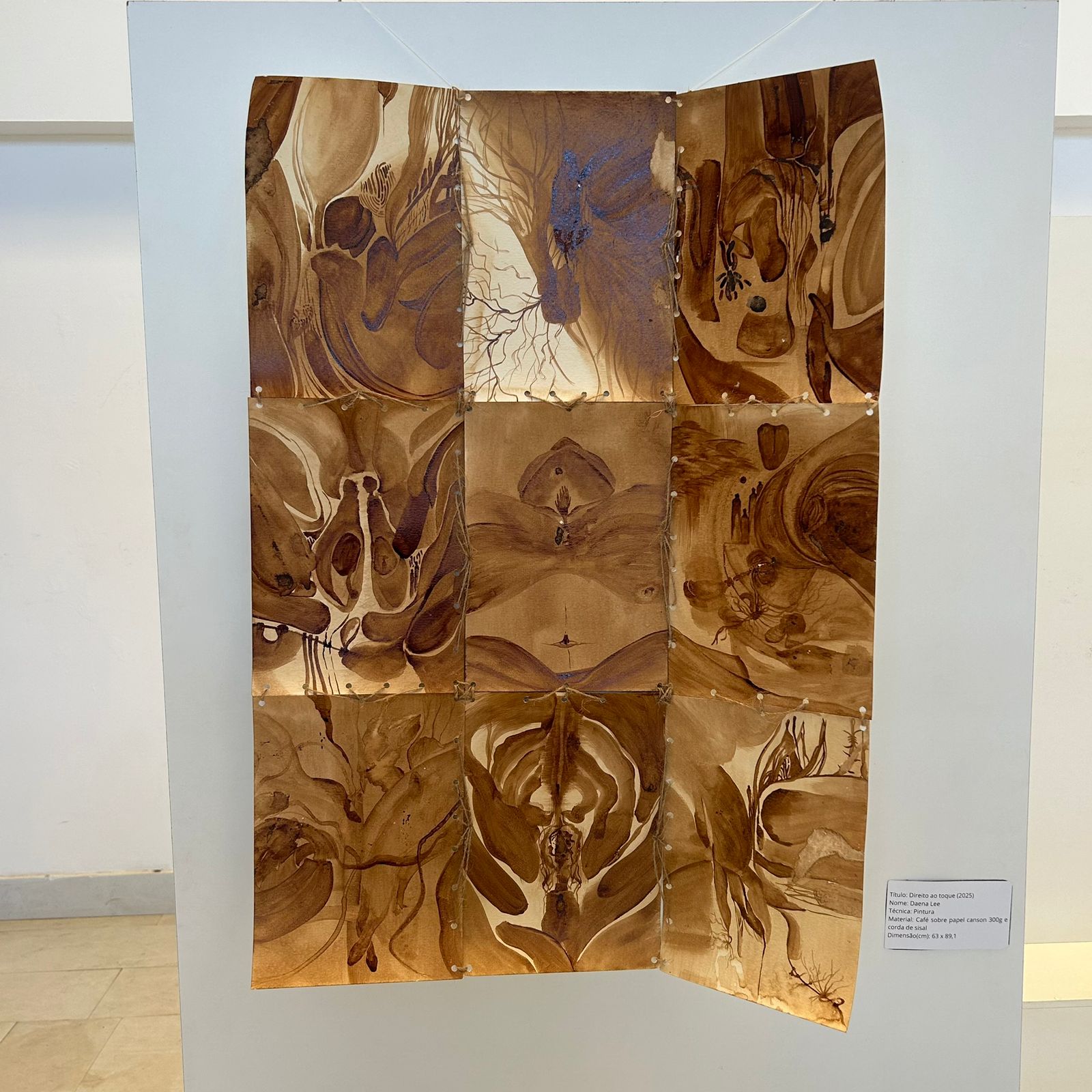 Fotografia de uma mesa expositora com tampa de vidro. Alguns livretos e colagens coloridas com títulos estão dispostas lado a lado, sobre algumas palhas da costa. Em uma, há a pintura de três pessoas em um fundo esbranquiçado, com uma flor amarela na parte superior. A segunda é um rosto de pele amarela, olhos alaranjados circulares, uma letra U no lugar do nariz e lábios abertos com os dentes à mostra. Ao lado, uma capa com fundo roxo e desenho em preto, dos olhos de uma pessoa com sobrancelhas arqueadas. Na parte de cima outra de capa lilás com desenho sinuoso de um corpo humano com os braços curvilíneos. Já na capa acima uma ilustração de prédios e o título em grafite “A cidade não era como hoje”. Ao lado, uma capa com colagens e estamparia colorida, abaixo, sobre um de capa lilás uma caderneta de capa vermelha e com o desenho da bandeira do Brasil no fecho. 
