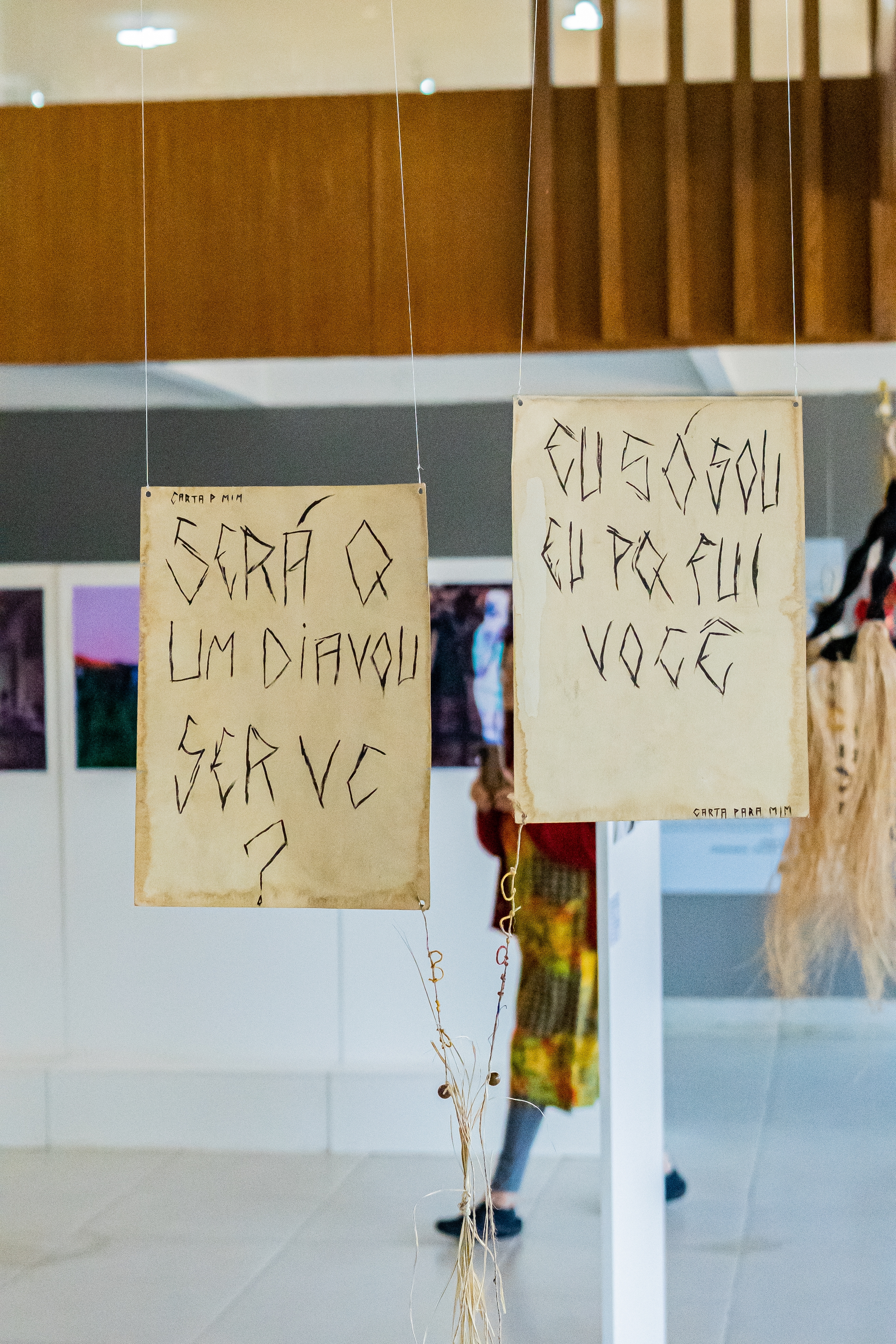 Fotografia de uma instalação. Duas placas em tons de papéis amarelados suspensas por fios, tem linhas pretas retas e estilizadas com frases. A da esquerda diz: “Será que um dia vou ser você?” Na parte superior, “Carta para mim”. A da direita, “ eu só sou eu porque fui você ?” Na parte inferior, “ Carta para mim”. Ao fundo, outras instalações e fotos. 
