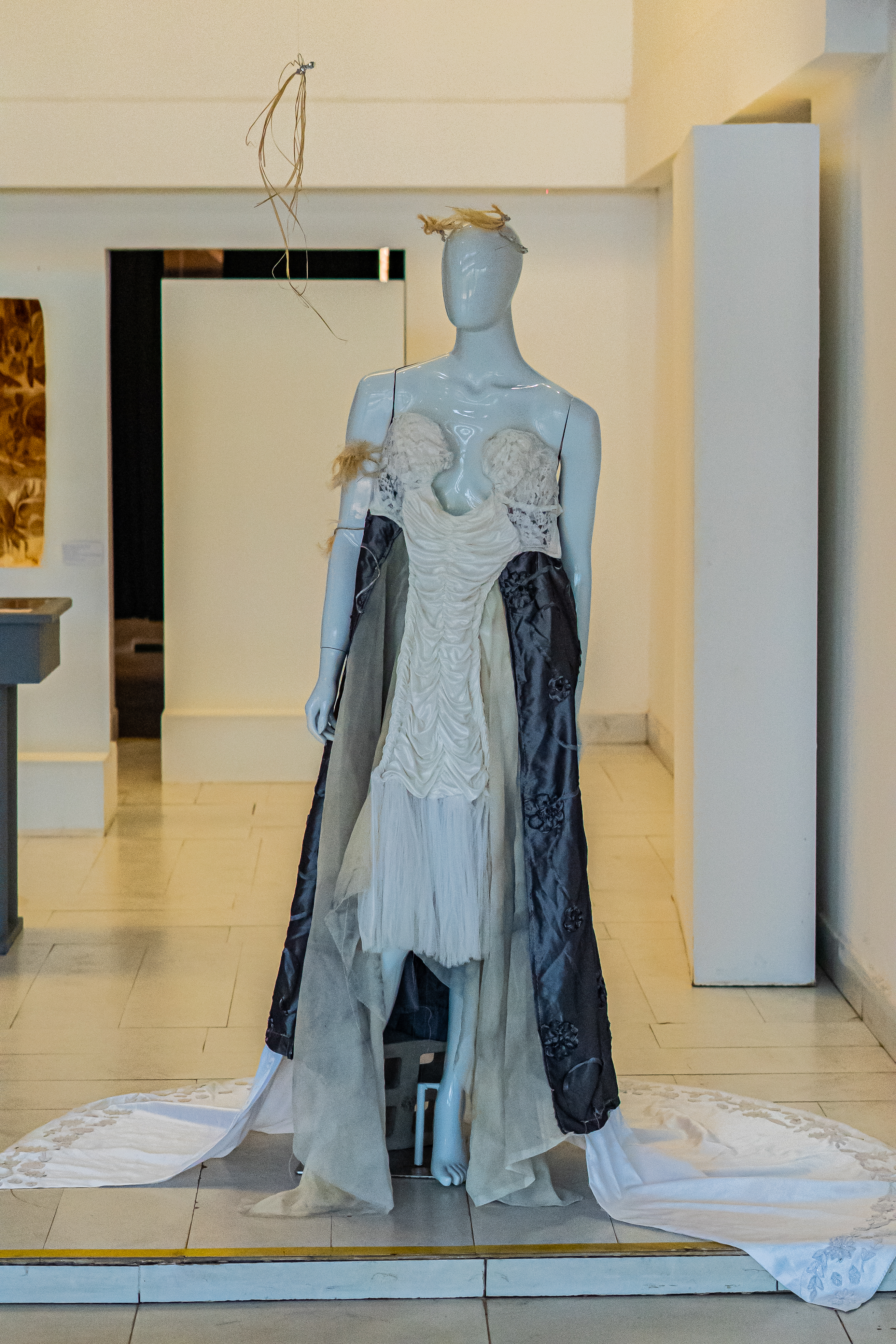Fotografia de um manequim de vestido em tons de bege e chumbo. Em uma sala com paredes brancas, o manequim usa na cabeça, uma tiara feita de palha e arame. No  braço direito, dois adornos também de palha. Está com um vestido sem alças com o busto bordado em creme, o mesmo tom de cor desce em um ponto plissado até um palmo antes do joelho. Abaixo, um pequeno caimento em tule. O saiote do vestido é longo, um tecido cor chumbo metálico, com bordados de flores e uma fenda na frente. Por baixo, uma longa cauda branca com bordado nas pontas.
