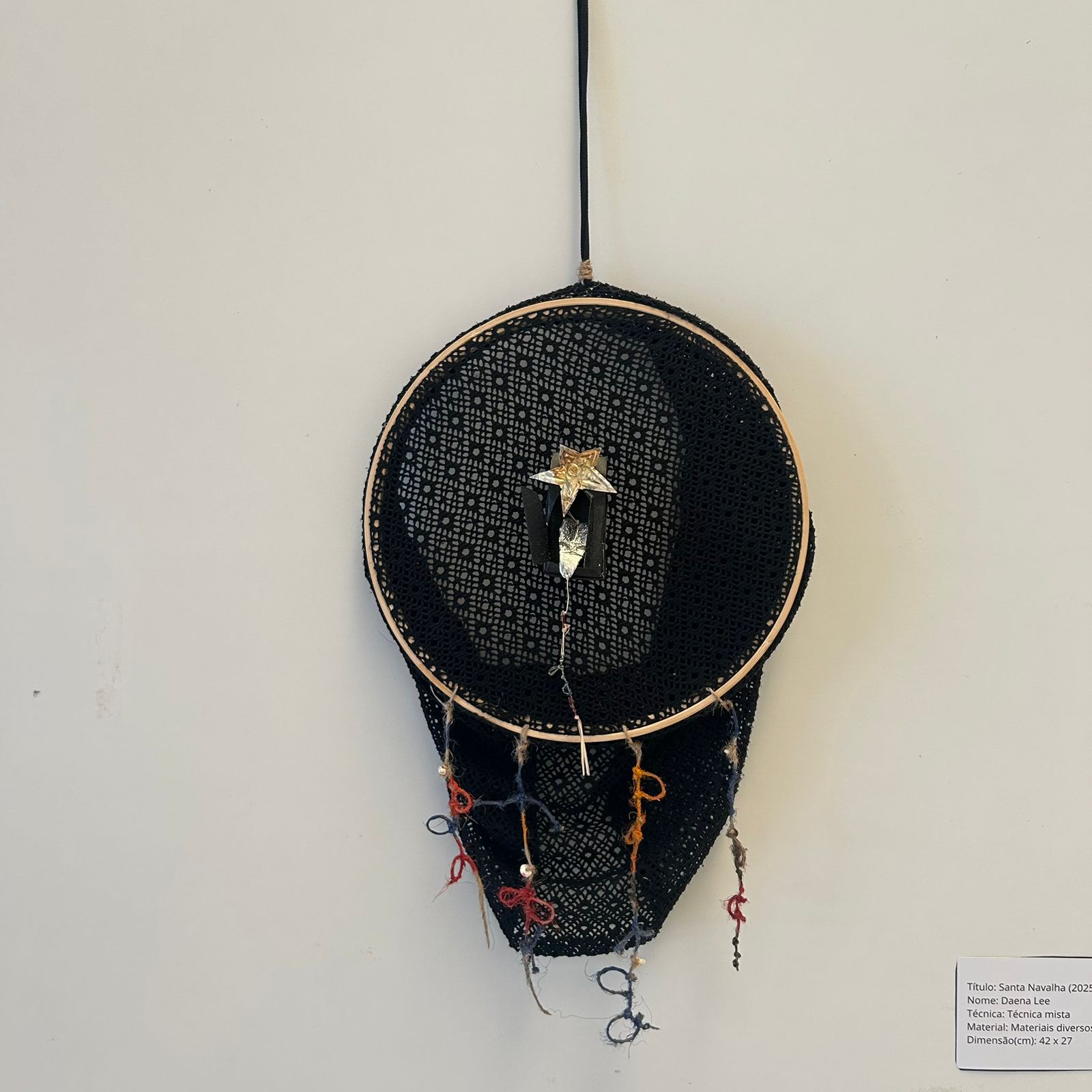 Fotografia de uma obra pendurada em uma parede branca. Um fio preto está preso em um aro que sustenta uma renda preta. No centro da peça, há uma estrela dourada em relevo, presa sobre uma peça prateada que se projeta para fora. Do aro inferior do bastidor, pendem fios coloridos soltos: vermelho, azul, laranja e bege amarrados em pequenos nós, alguns finalizados com miçangas brancas.
