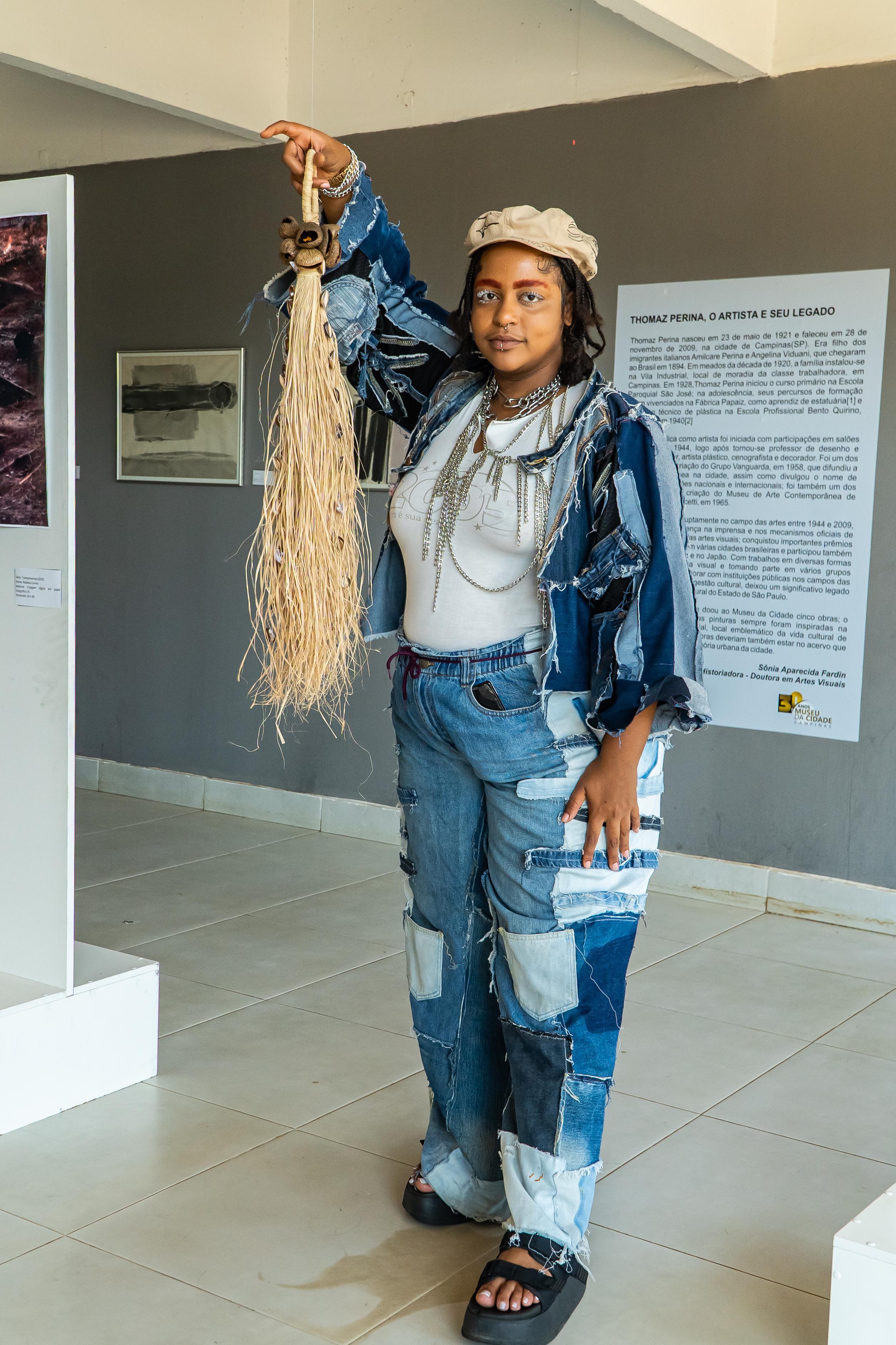 Fotografia de uma pessoa segurando uma obra. Em uma sala com exposições ao fundo e paredes cinzas e detalhes em branco. A pessoa  é negra de pele clara com tranças curtas até a altura do ombro, usa boina bege, camiseta branca, jaqueta e calça jeans de retalhos.; a pessoa está de sobrancelhas pintadas de laranja e os cílios brancos. Usa diversos colares de prata e esboça um leve sorriso. A obra é composta por palha da costa trançada em uma alça, na base um adorno de coquinhos e semente marrom. A - alça se abre em fios separados com um caimento longo e volumoso, entre as palhas da costa fios de búzios nigerianos abertos. 
