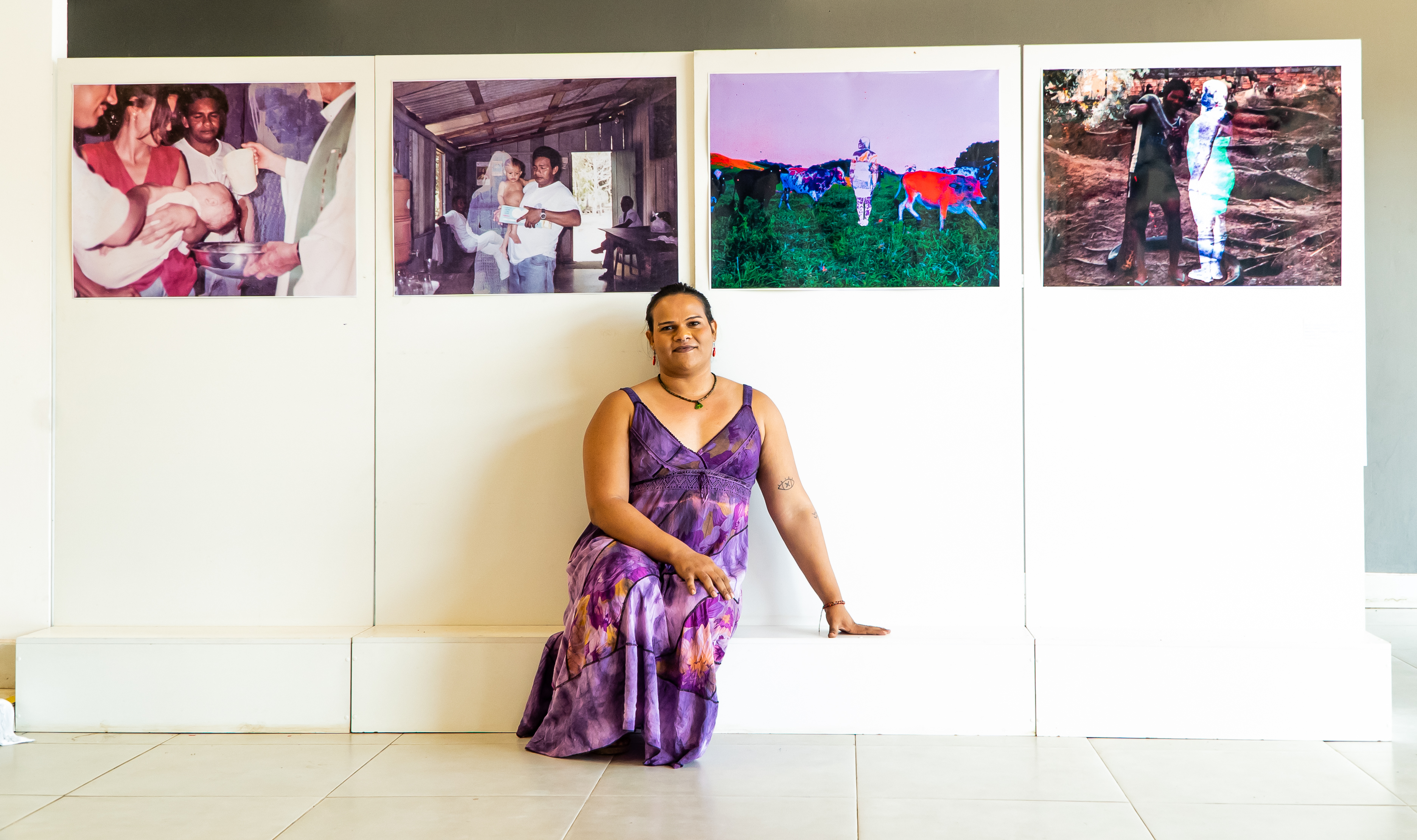  Fotografia. Uma pessoa sentada em um banco. Ao fundo, uma parede branca com quatro fotografias coloridas. A pessoa é negra de pele clara, tem olhos pequenos, sobrancelhas finas e arredondadas. O nariz núbio e lábios grossos, usa maquiagem escura. Está de vestido de alças, longo e roxo. As fotos mostram cenas de pessoas em momentos cotidianos e uma imagem de vacas no campo. Na primeira foto, uma criança de pele clara é batizada. Um homem e uma mulher seguram um bebê, que está deitado de peito para cima, um padre segura uma bacia de metal embaixo da cabeça da criança e segura um jarro com água, ao fundo um homem negro observa.  Na segunda foto, o mesmo homem carrega um bebê de colo em uma casa de madeira. O homem negro tem pele retinta, segura o bebê no colo, e uma embalagem. do lado esquerdo a silhueta em tons de branco de uma mulher de cabelo longo; ao fundo duas pessoas observam mais de trás. Na terceira imagem, foto de um pasto com uma silhueta. Cinco vacas caminham no pasto, ao centro uma silhueta esbranquiçada de uma pessoa, ao fundo um monte laranja fluorescente e mesmo tom em uma vaca do lado direito. Na última foto, um homem segura uma cobra e sorri, junto a silhueta esbranquiçada de uma pessoa em um morro de terra com raízes . 

