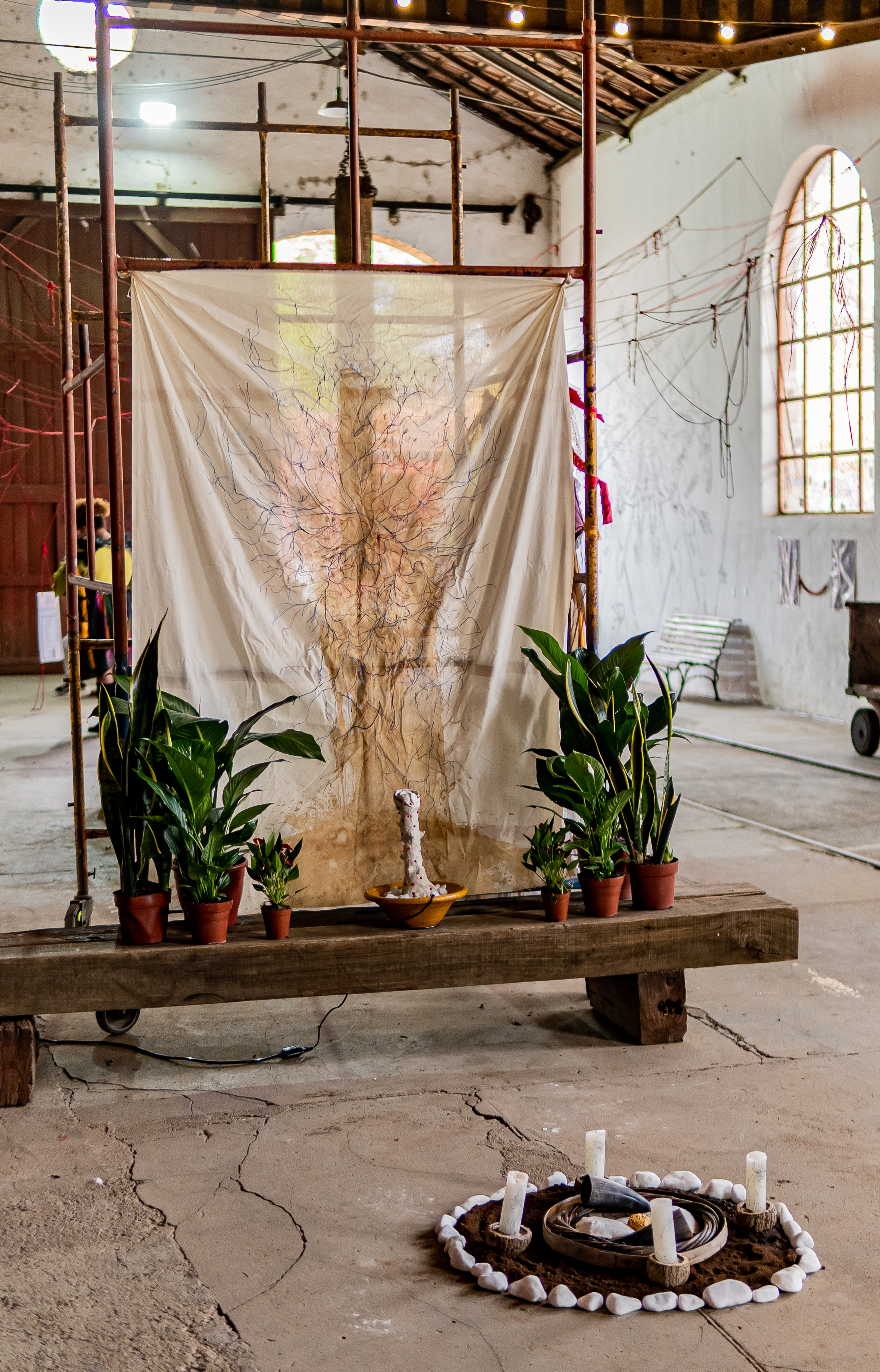 Fotografia de uma instalação em um espaço fechado com paredes brancas e janelas grandes. Em um andaime, um tecido branco esticado com um desenho disforme de um tronco de árvore grosso, feito com uma mancha amarelada.  no lugar da copa, ramificações em vermelho e azul, partindo do centro para as laterais.  Embaixo do andaime, está um banco de madeira, sobre o móvel, vasos marrons com plantas do lado esquerdo e do lado direito. São quatro de cada lado com as mesmas plantas: Espada de Iansã, lírio da paz, folhas de figo. Ao centro do banco, um alguidar marrom médio, dentro um objeto branco longo fálico, com pequenas esferas vermelhas grudadas na base do alguidar pedras brancas. 
