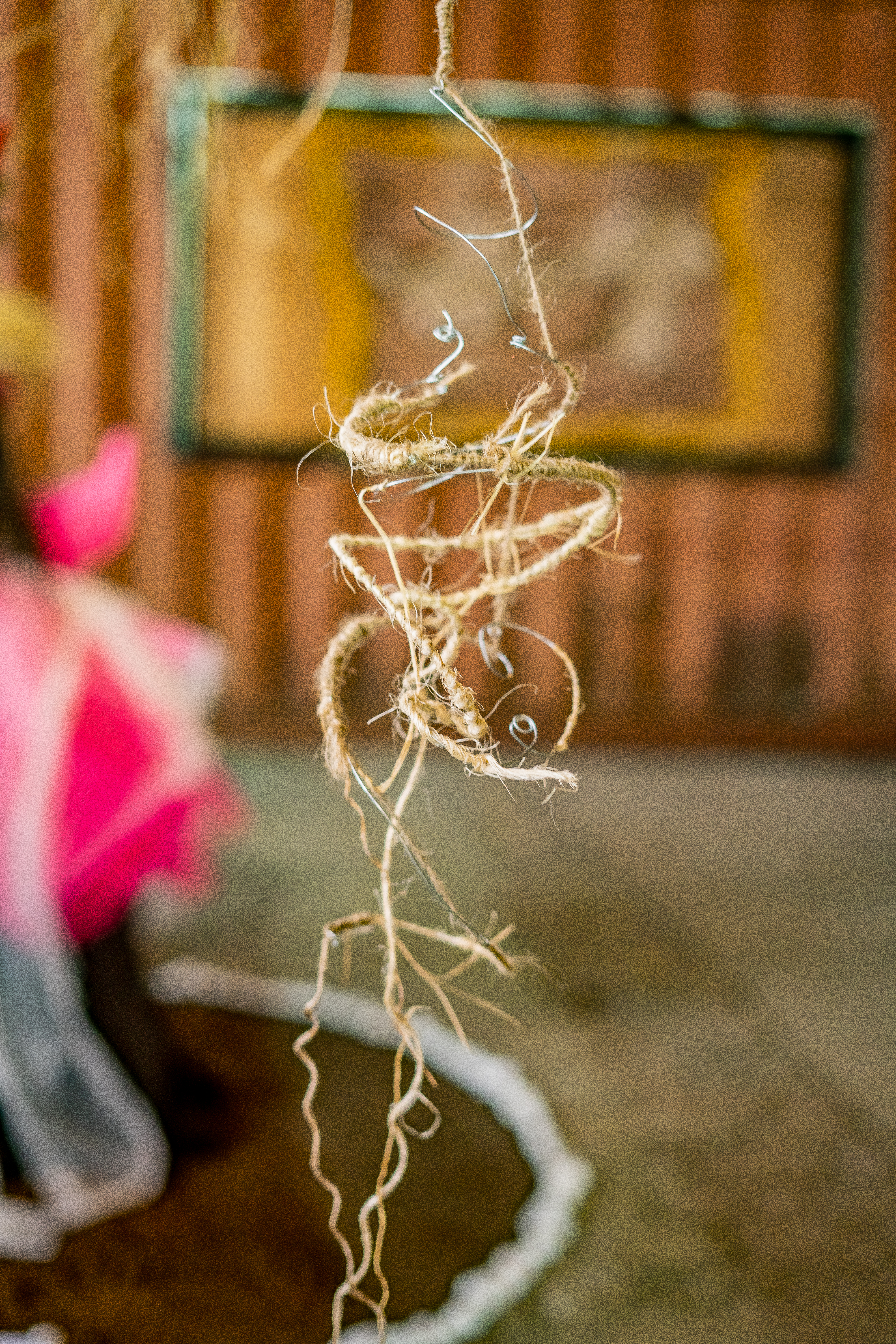 Fotografia de uma instalação. Em uma sala, desfocada, a obra é um fio de palha da costa enrolado em um fio de aço. A palha e o fio se enovelam-se, ela desfia e se solta nas extremidades da peça.   
