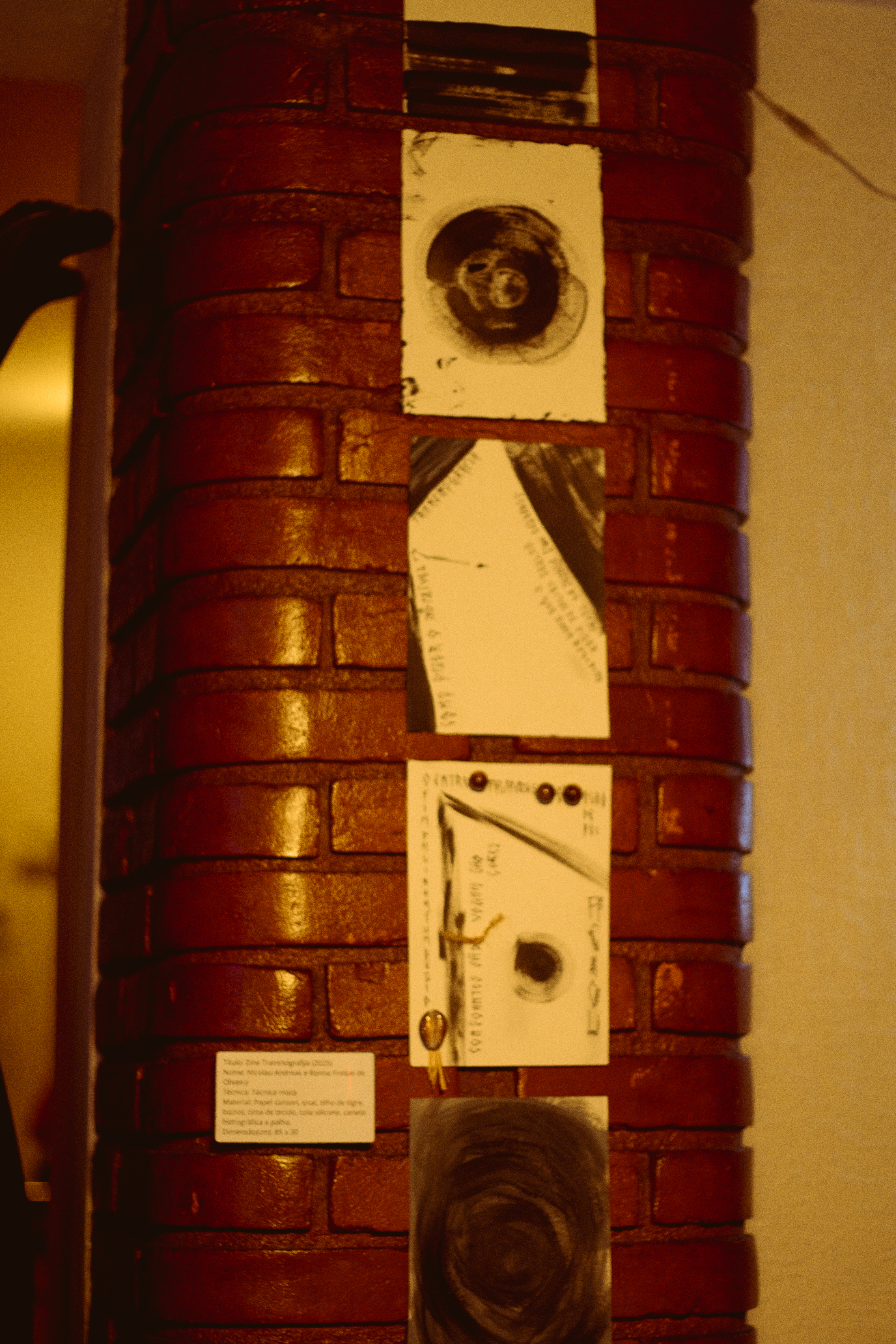 Foto de uma instalação. Em um pilar de tijolos amarelos, cinco quadros, formando uma linha vertical. Em cada um, figuras abstratas e textos ilegíveis. O primeiro, vista pela metade, é um desenho de riscos horizontais. No segundo, há uma concentração no meio da folha de desenhos circulares. No terceiro, há pinturas nas pontas superiores e na inferior direita, alguns textos escritos também nas bordas. No penúltimo, duas linhas constam textos, uma vertical longa e uma diagonal. No último, visto pela metade, há um borrão também circular. 

