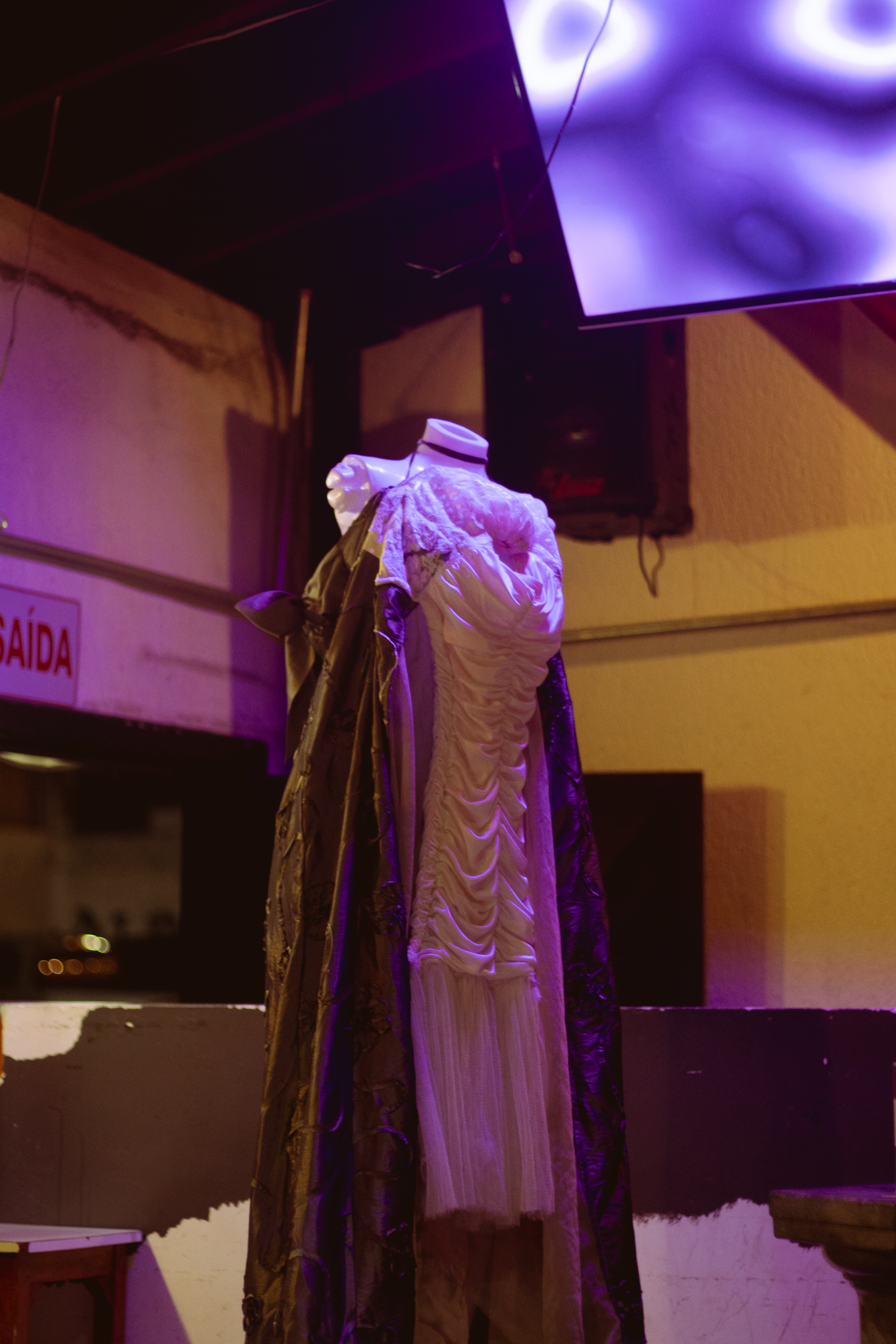 Fotografia de um manequim com vestido em tons de bege e chumbo. Em uma sala com paredes brancas, o manequim usa um vestido sem alças com o busto bordado em creme. O mesmo tom de cor desce em um ponto plissado até um palmo antes do joelho, abaixo um pequeno caimento em tule. O saiote do vestido é longo, um tecido cor chumbo metálico tem bordados de flores e uma fenda na frente. Por baixo, uma longa cauda branca com bordado nas pontas. Na cabeça do manequim, uma tiara feita de palha e arame, e no braço direito dois adornos também de palha.
