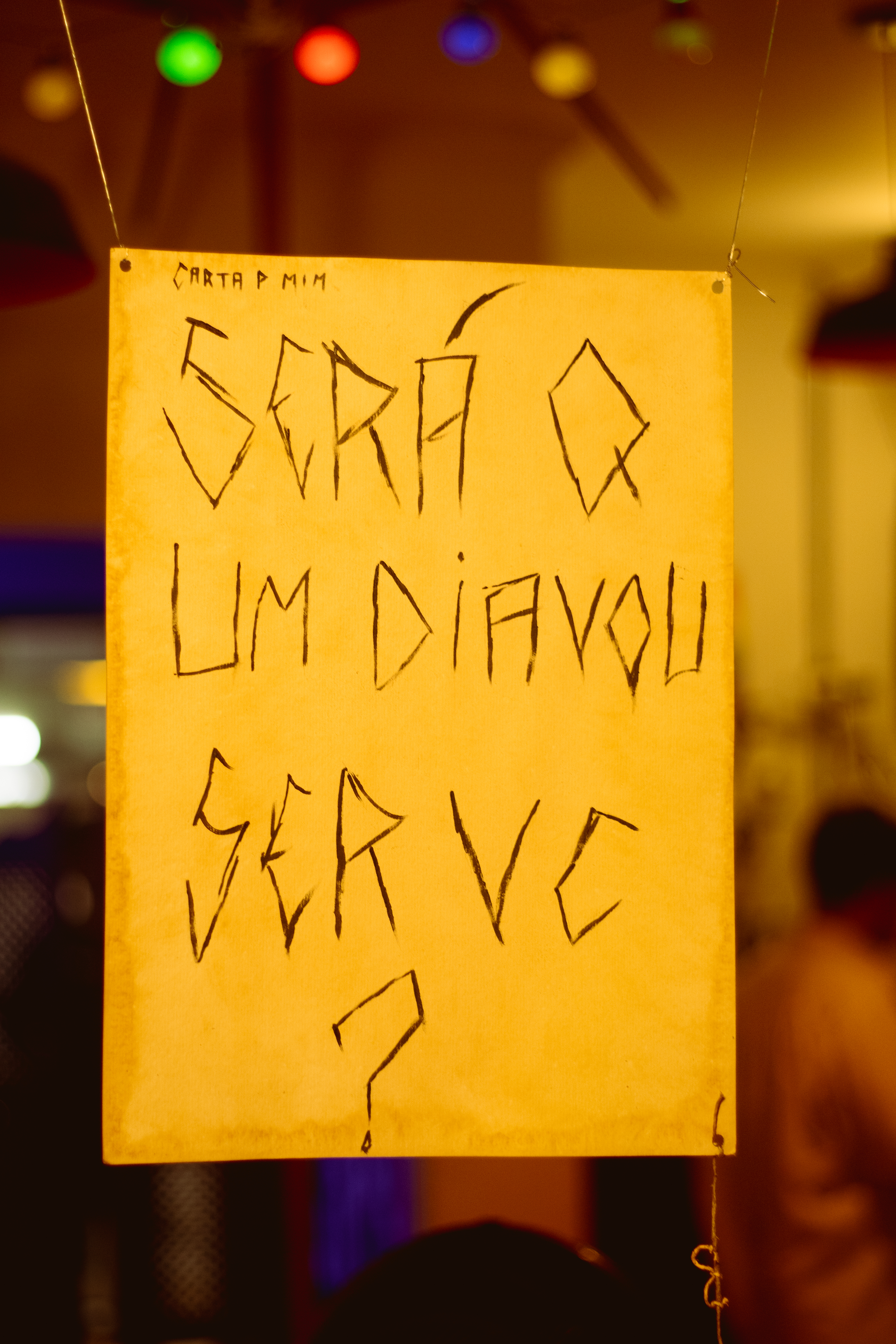 Fotografia de uma instalação. Uma placa em tons de papel amarelos suspenso por fios, em letras grafitadas a pergunta: “Será que um dia vou ser você?” A palavra “que” é composta só pela letra Q e palavra “você” pelas consoantes V e C.
