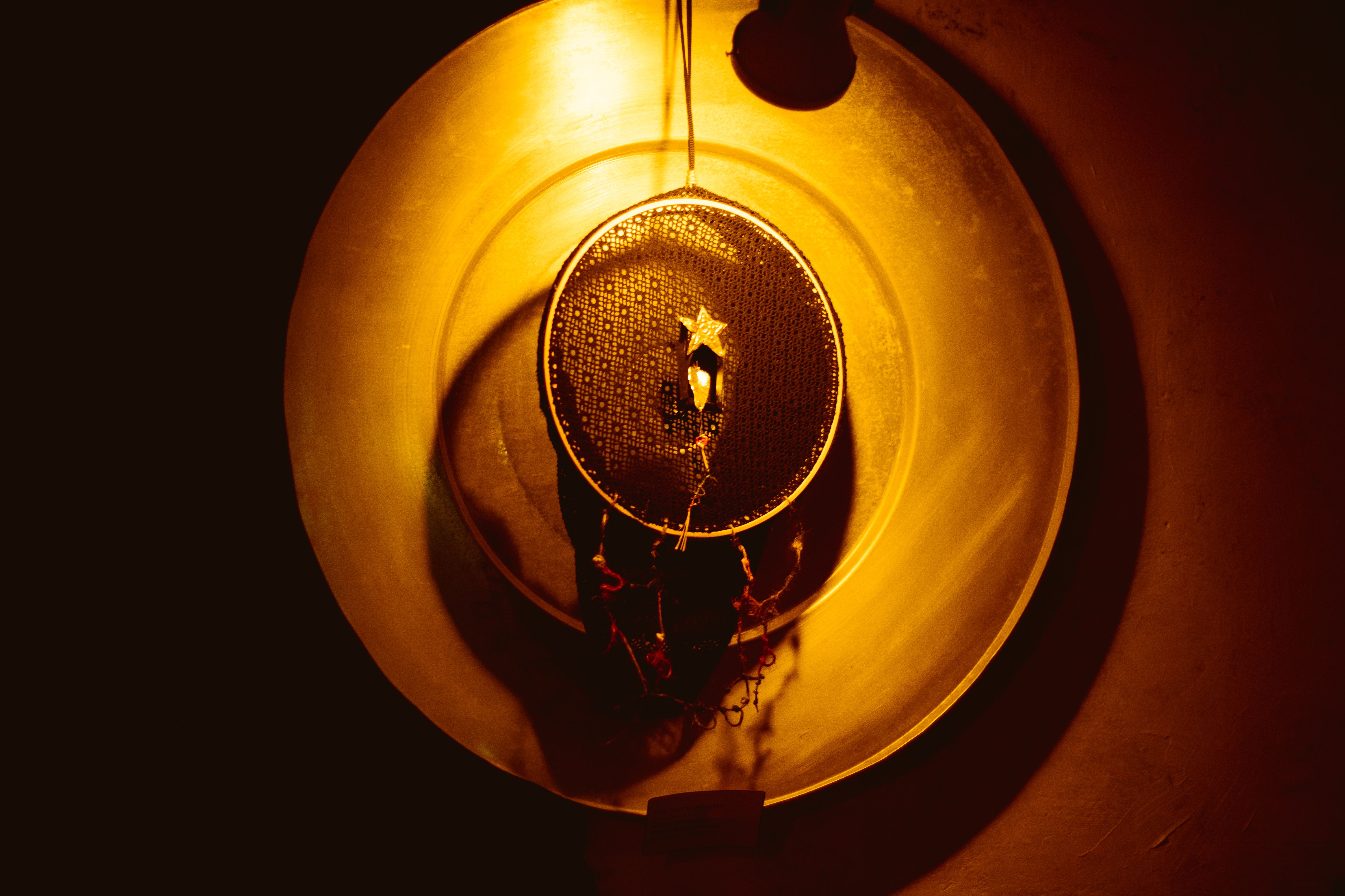 Fotografia de uma obra pendurada em uma parede branca.  Ela está sobre um prato raso e tons de bronze. A foto tem iluminação amarelada. A obra é um fio preto está preso em um aro que sustenta uma renda preta. No centro da peça, há uma estrela dourada em relevo, presa sobre uma peça prateada que se projeta para fora, Do aro inferior do bastidor, pendem fios coloridos soltos: vermelho, azul, laranja e bege amarrados em pequenos nós, alguns finalizados com miçangas brancas.
