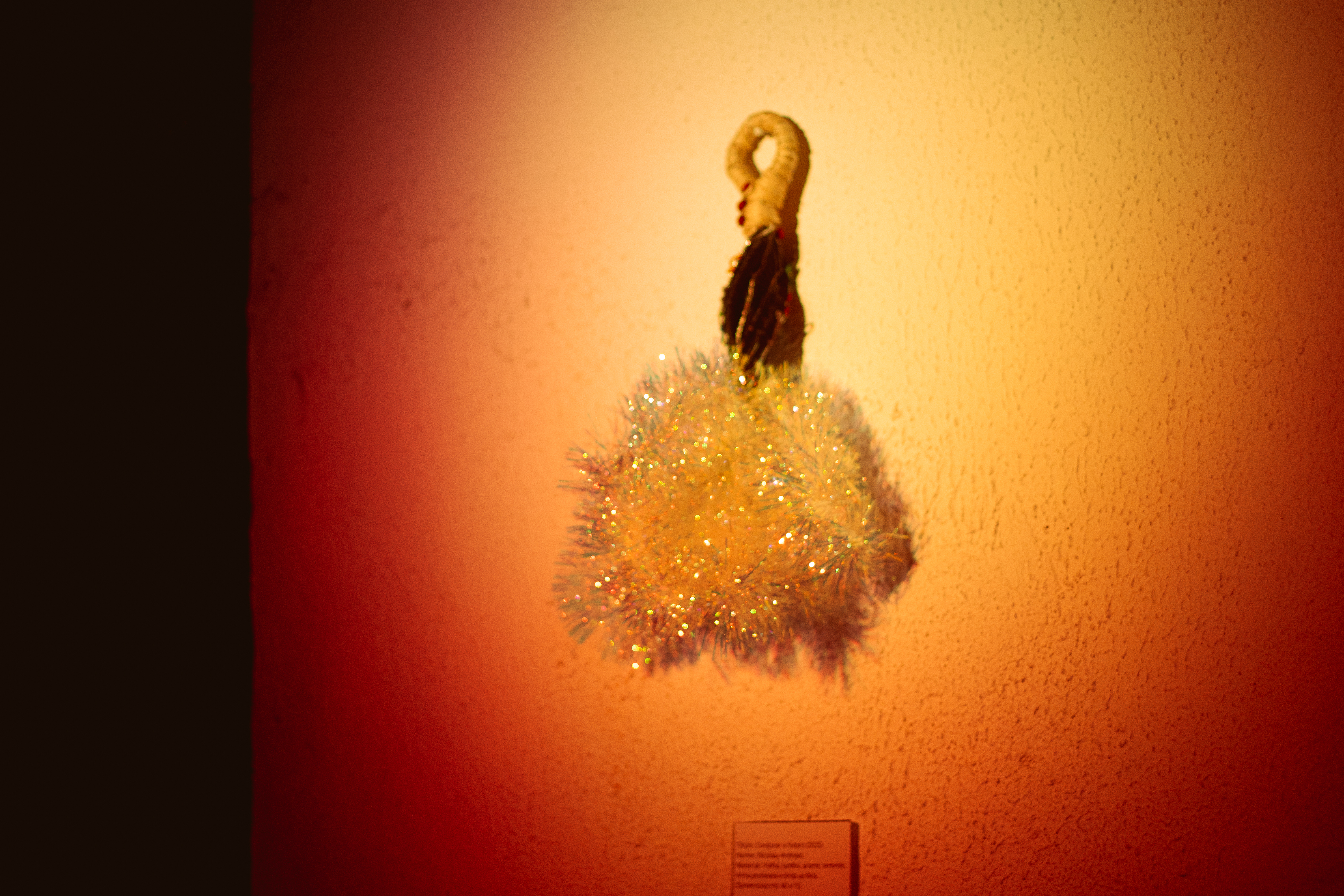 Fotografia de uma instalação. Pendurada em uma parede clara e iluminada, a pequena obra tem uma alça de palha com pequenas bolinhas vermelhas, na base um ondulado marrom e na ponta um festão em furta cor.